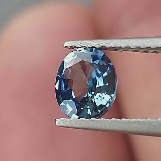 Blue-gray oval sapphire shown rotating in tweezers, between fingertips for scale, on a palm and on a white pad.