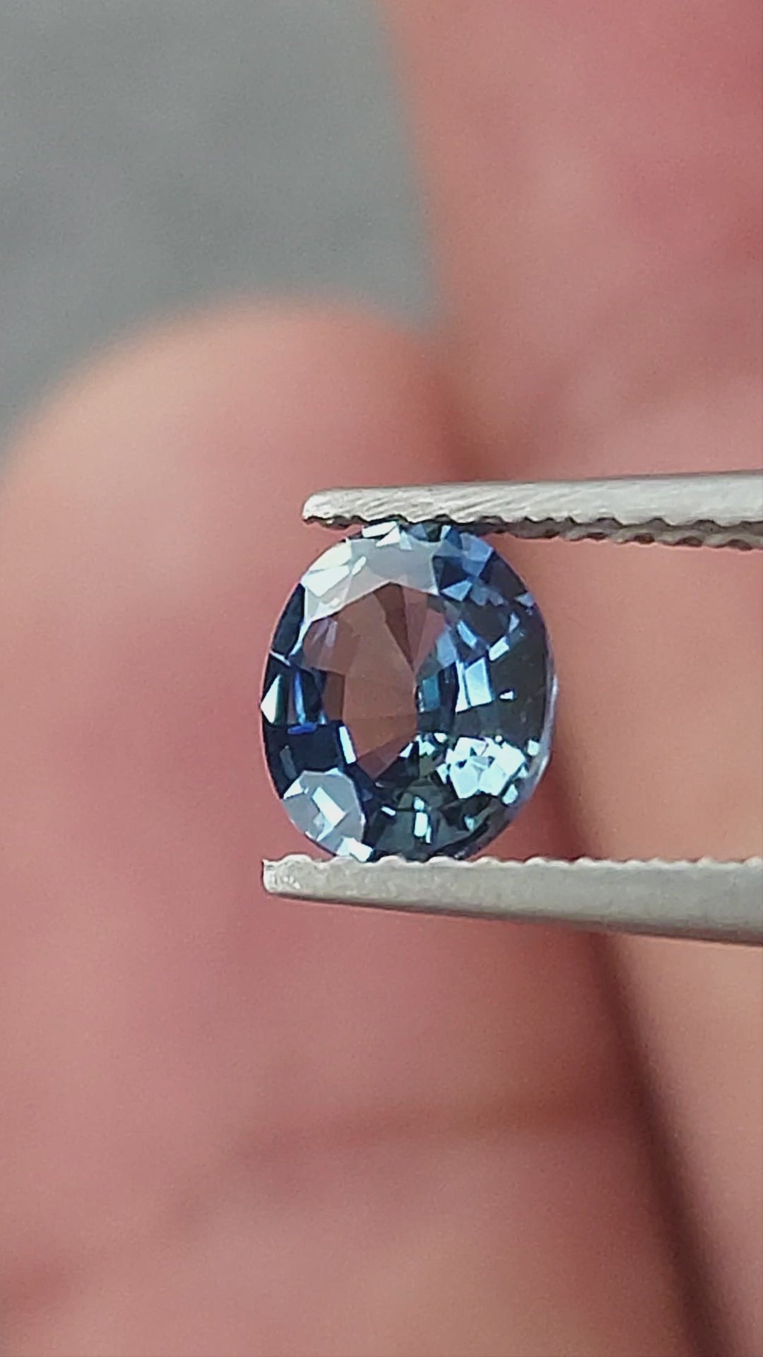 Blue-gray oval sapphire shown rotating in tweezers, between fingertips for scale, on a palm and on a white pad.