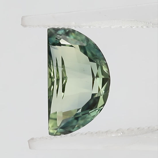 Video views of the half-moon parti sapphire in tweezers and on fingertips for scale, including its face-up appearance and checkerboard pavilion, with blue-green and pale yellow-green tones.