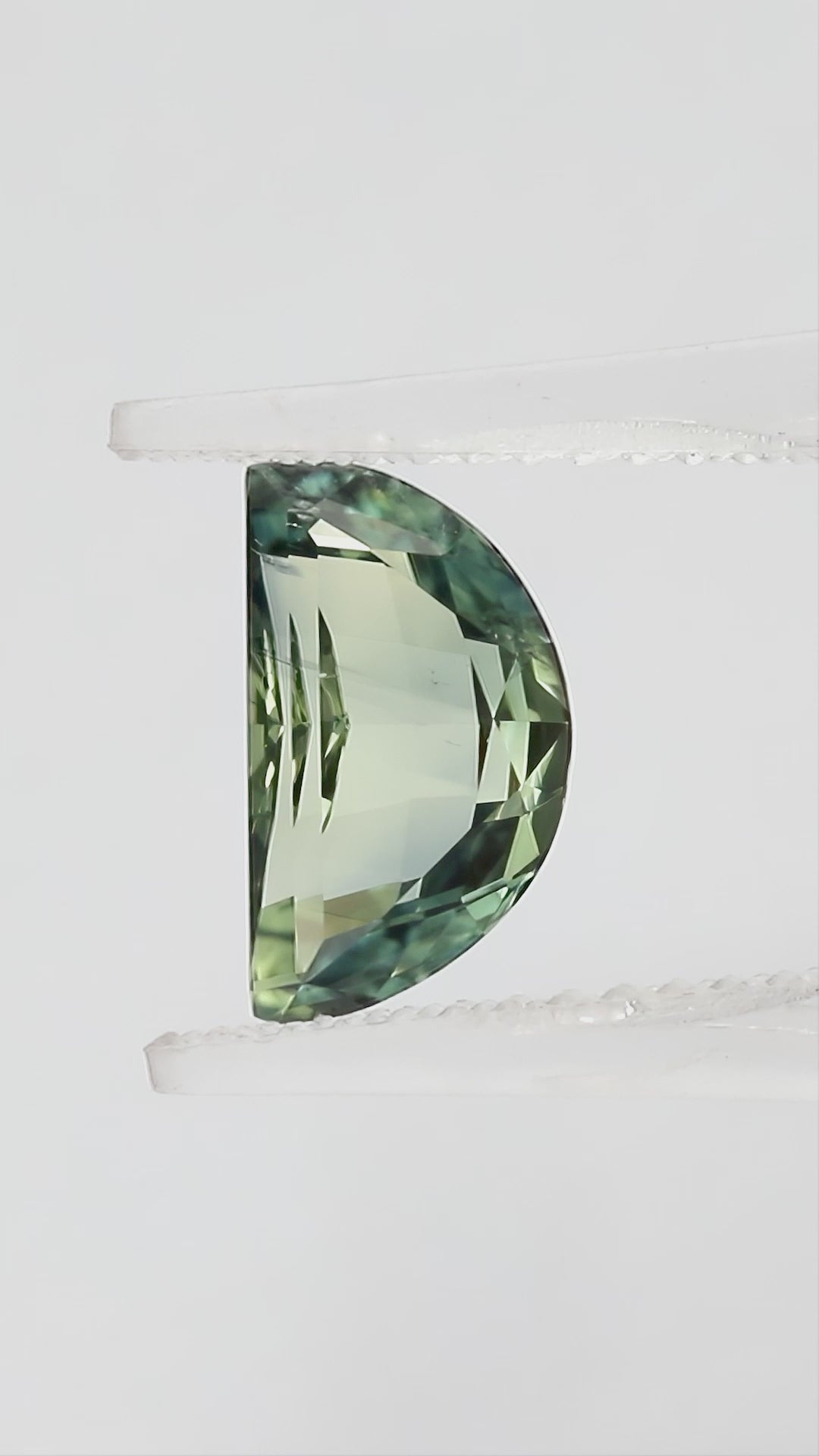 Video views of the half-moon parti sapphire in tweezers and on fingertips for scale, including its face-up appearance and checkerboard pavilion, with blue-green and pale yellow-green tones.