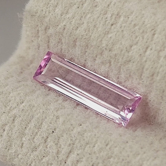 Pink baguette sapphire shown on a cream pad, between fingertips for scale and rotating in tweezers.