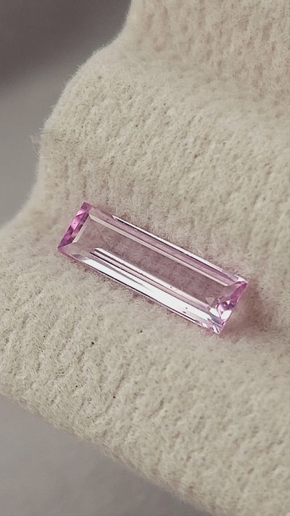 Pink baguette sapphire shown on a cream pad, between fingertips for scale and rotating in tweezers.