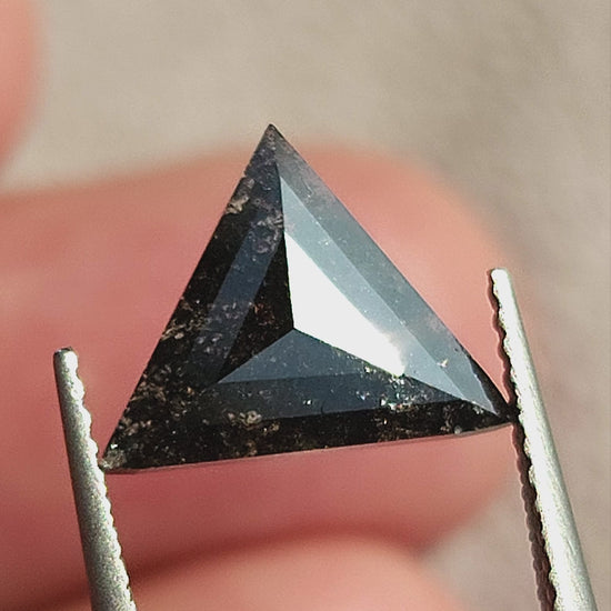 Triangular rose-cut salt and pepper diamond rotated in tweezers, showing crown, shallow profile and inclusions, followed by a view on fingertips for scale.