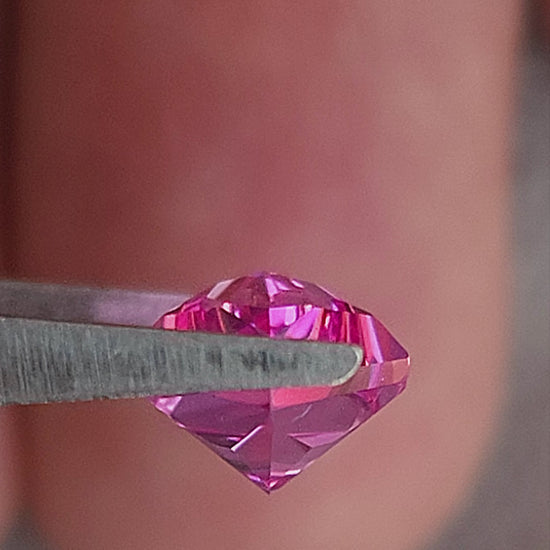 Hot pink geometric kite-cut sapphire tilted in tweezers and shown on fingers for scale.