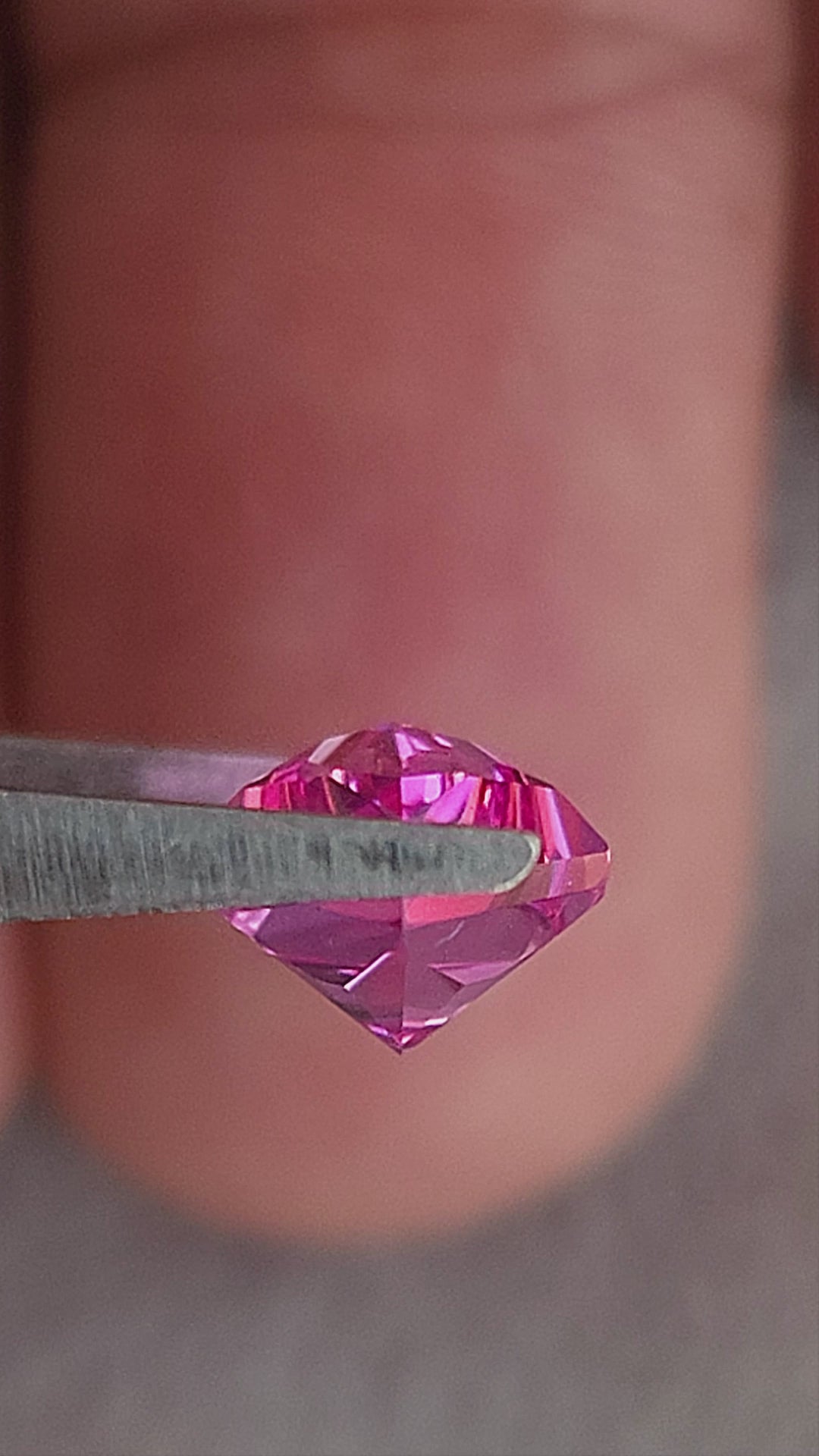 Hot pink geometric kite-cut sapphire tilted in tweezers and shown on fingers for scale.