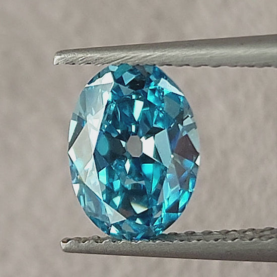 Blue oval diamond rotating in tweezers and then between fingertips, showing its broad facets and size.