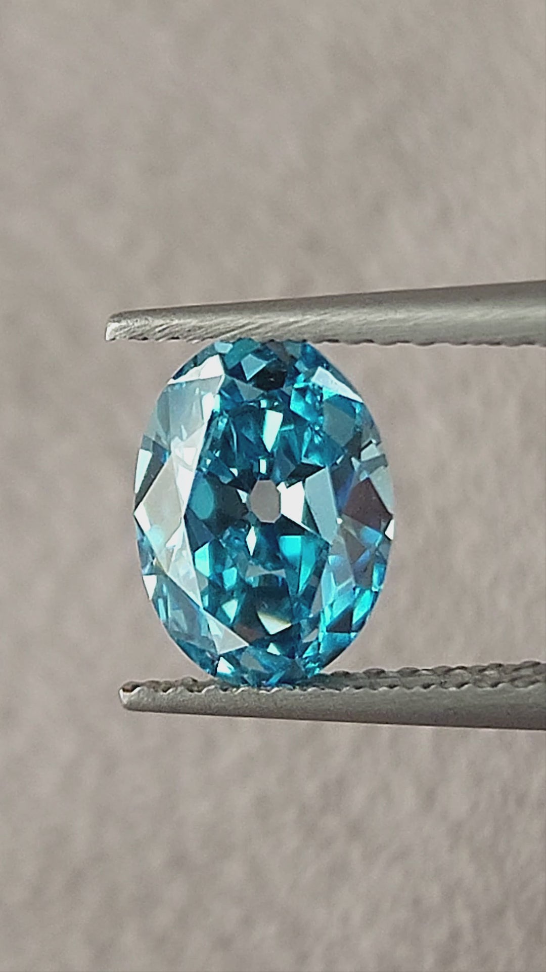 Blue oval diamond rotating in tweezers and then between fingertips, showing its broad facets and size.