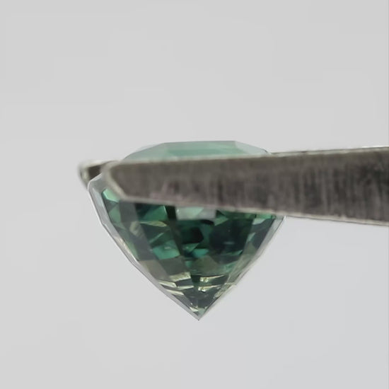 Teal-green oval sapphire turning in tweezers and resting between fingertips for scale.