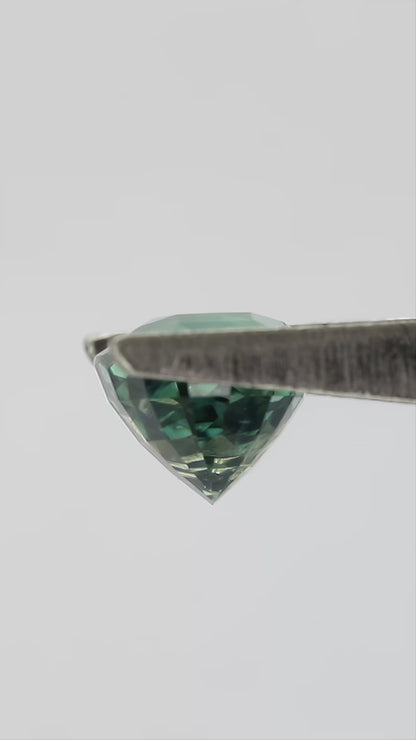 Teal-green oval sapphire turning in tweezers and resting between fingertips for scale.