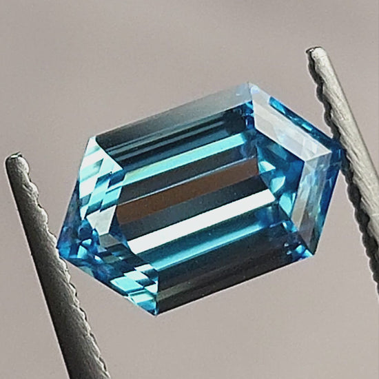Video of the blue elongated hexagon step-cut diamond in tweezers and on a white surface, showing pale and dark central reflections at different angles.