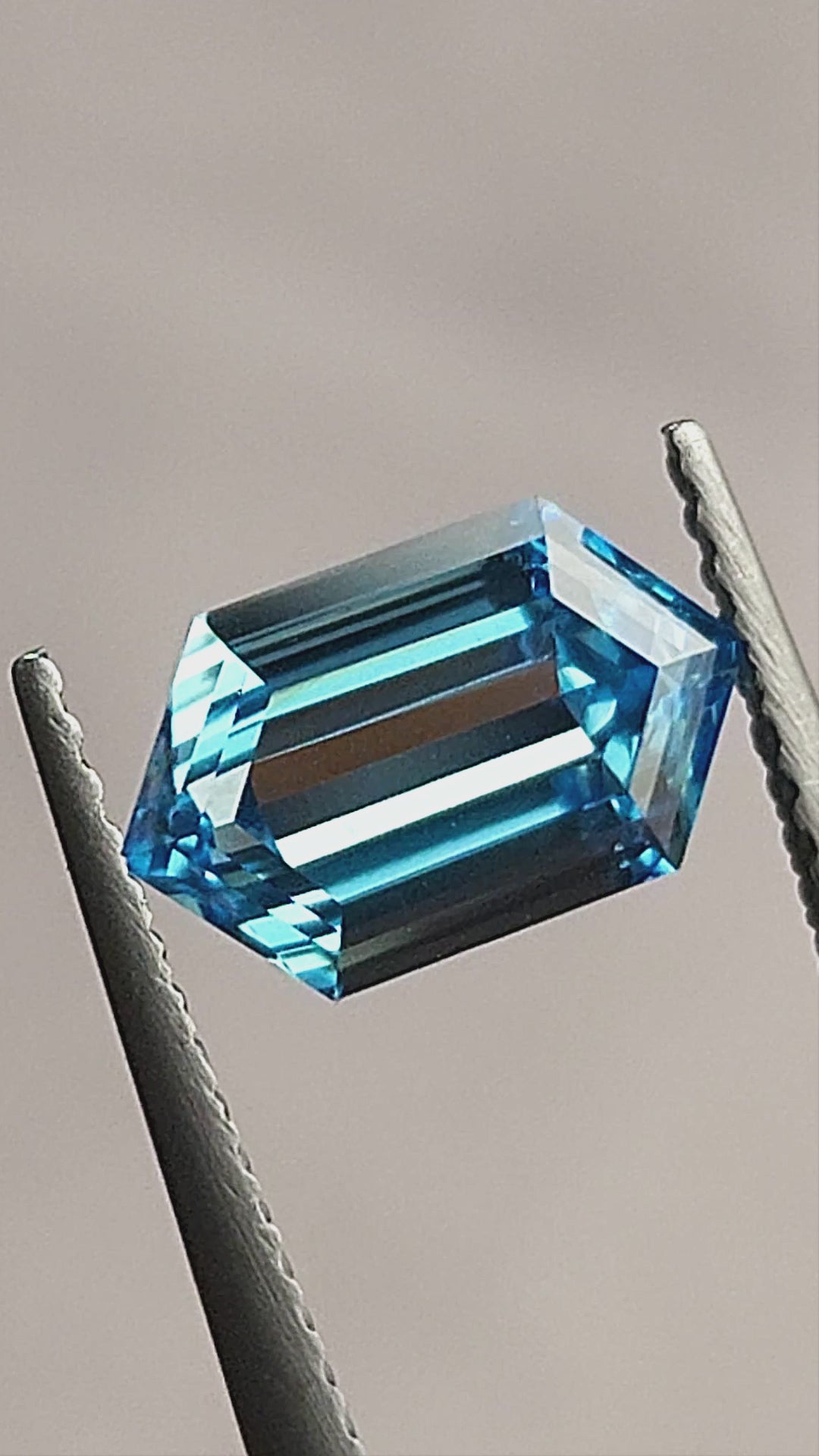 Video of the blue elongated hexagon step-cut diamond in tweezers and on a white surface, showing pale and dark central reflections at different angles.
