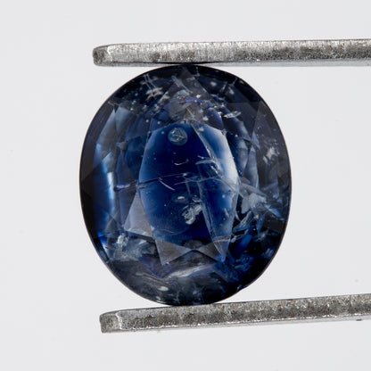 Face-up view of the oval blue sapphire in tweezers, showing its outline, table and visible inclusions.