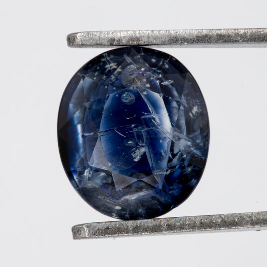 Face-up view of the oval blue sapphire in tweezers, showing its outline, table and visible inclusions.