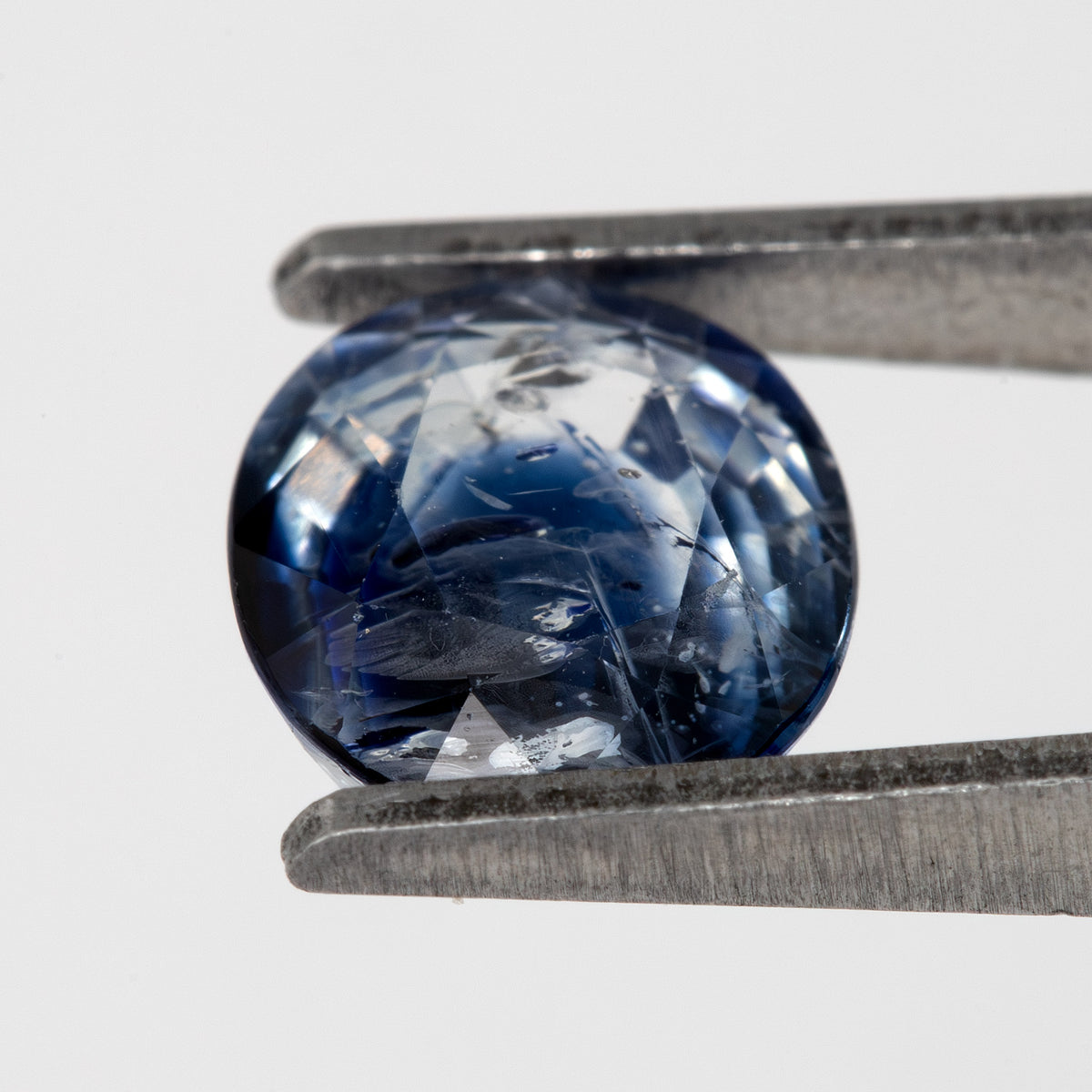 Tilted crown-side view of the oval blue sapphire in tweezers, showing a different pattern of facet reflections.