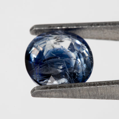 Tilted crown-side view of the oval blue sapphire in tweezers, showing a different pattern of facet reflections.