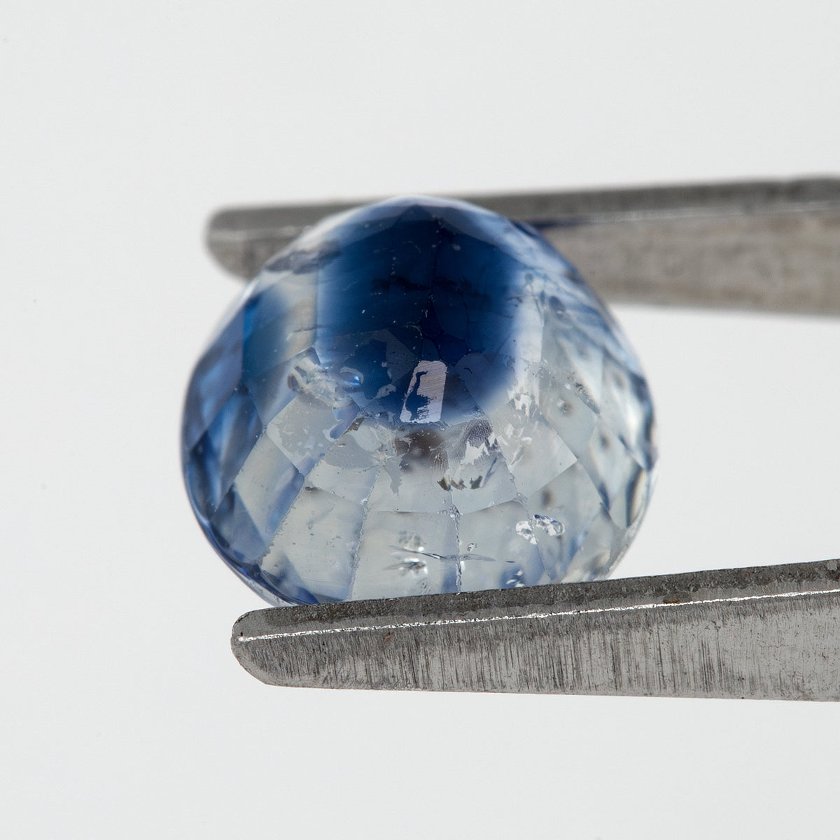 Tilted back view of the oval blue sapphire in tweezers, showing its pavilion facets.