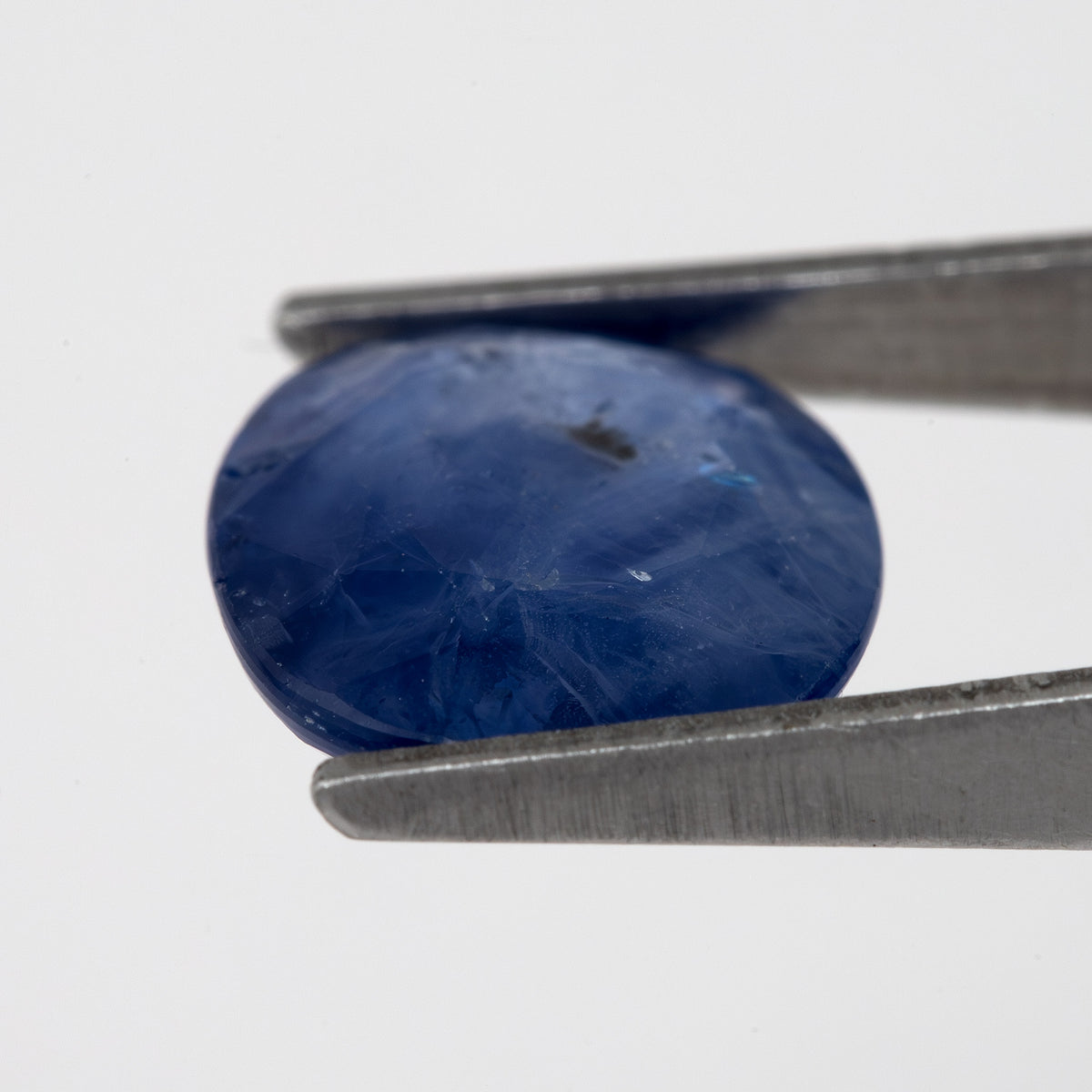 Tilted crown-side view of the oval blue sapphire in tweezers, showing a different pattern of facet reflections.