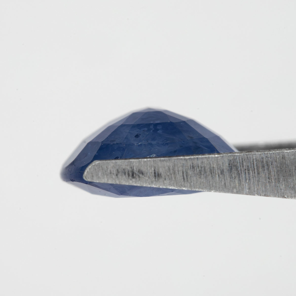 Side profile of the oval blue sapphire held in tweezers, showing crown and pavilion depth.