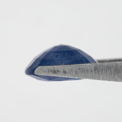 Side profile of the oval blue sapphire held in tweezers, showing crown and pavilion depth.