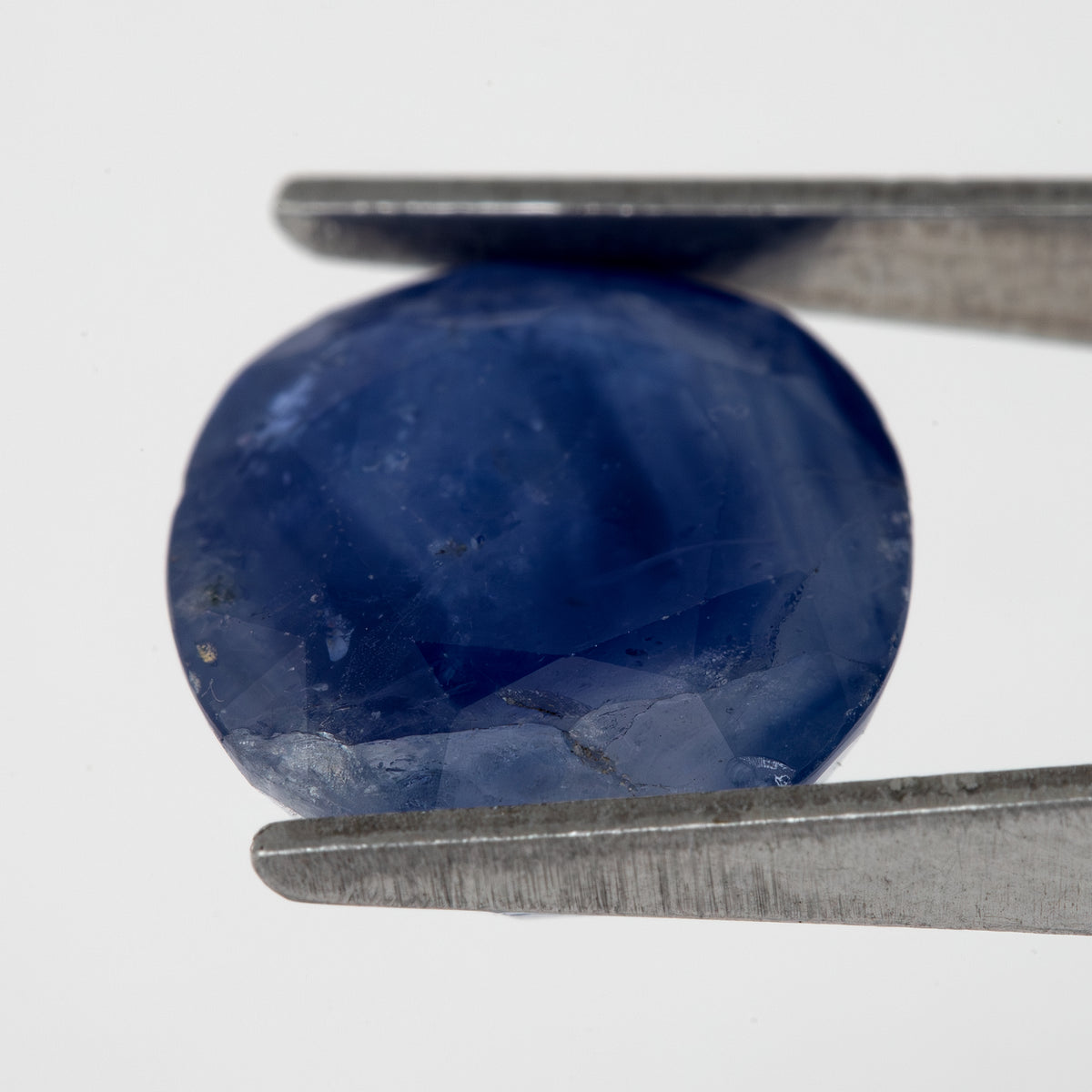 Tilted crown-side view of the oval blue sapphire in tweezers, showing a different pattern of facet reflections.