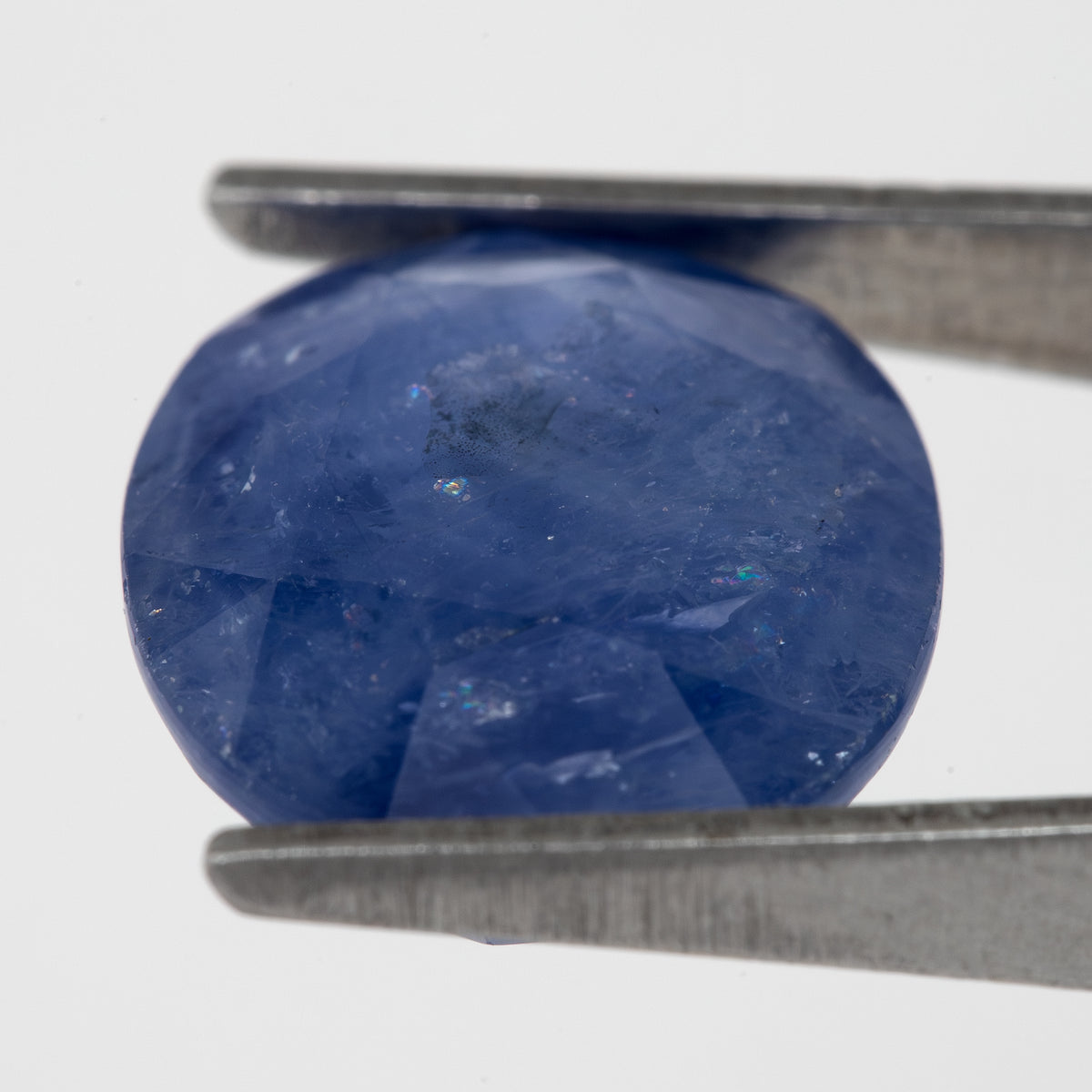 Tilted crown-side view of the oval blue sapphire in tweezers, showing a different pattern of facet reflections.