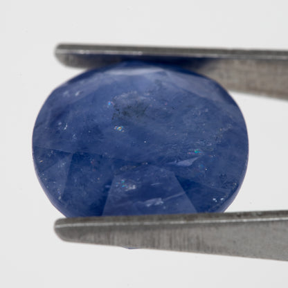 Tilted crown-side view of the oval blue sapphire in tweezers, showing a different pattern of facet reflections.