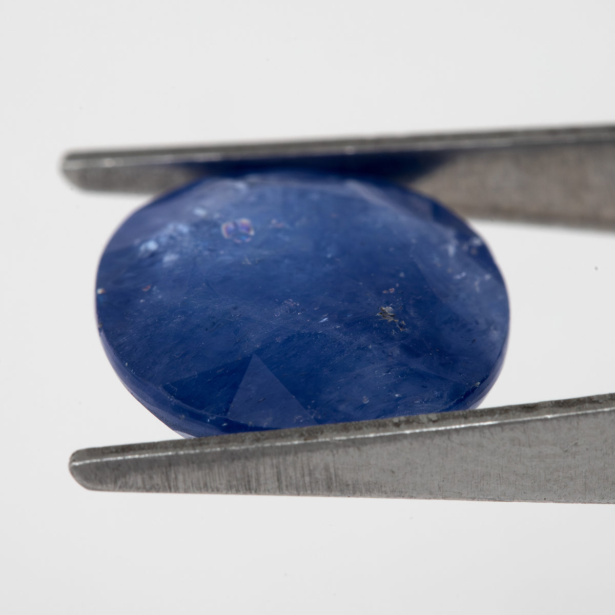 Tilted crown-side view of the oval blue sapphire in tweezers, showing a different pattern of facet reflections.