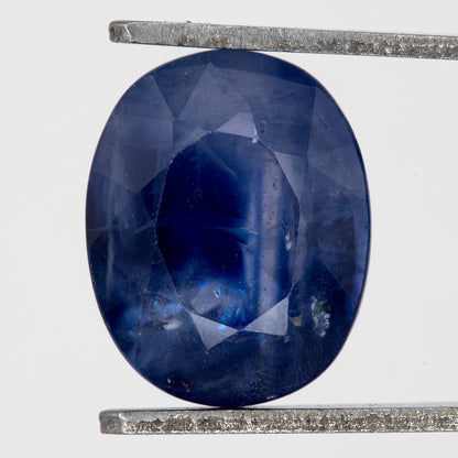 Face-up view of the oval blue sapphire in tweezers, showing its outline, table and visible inclusions.