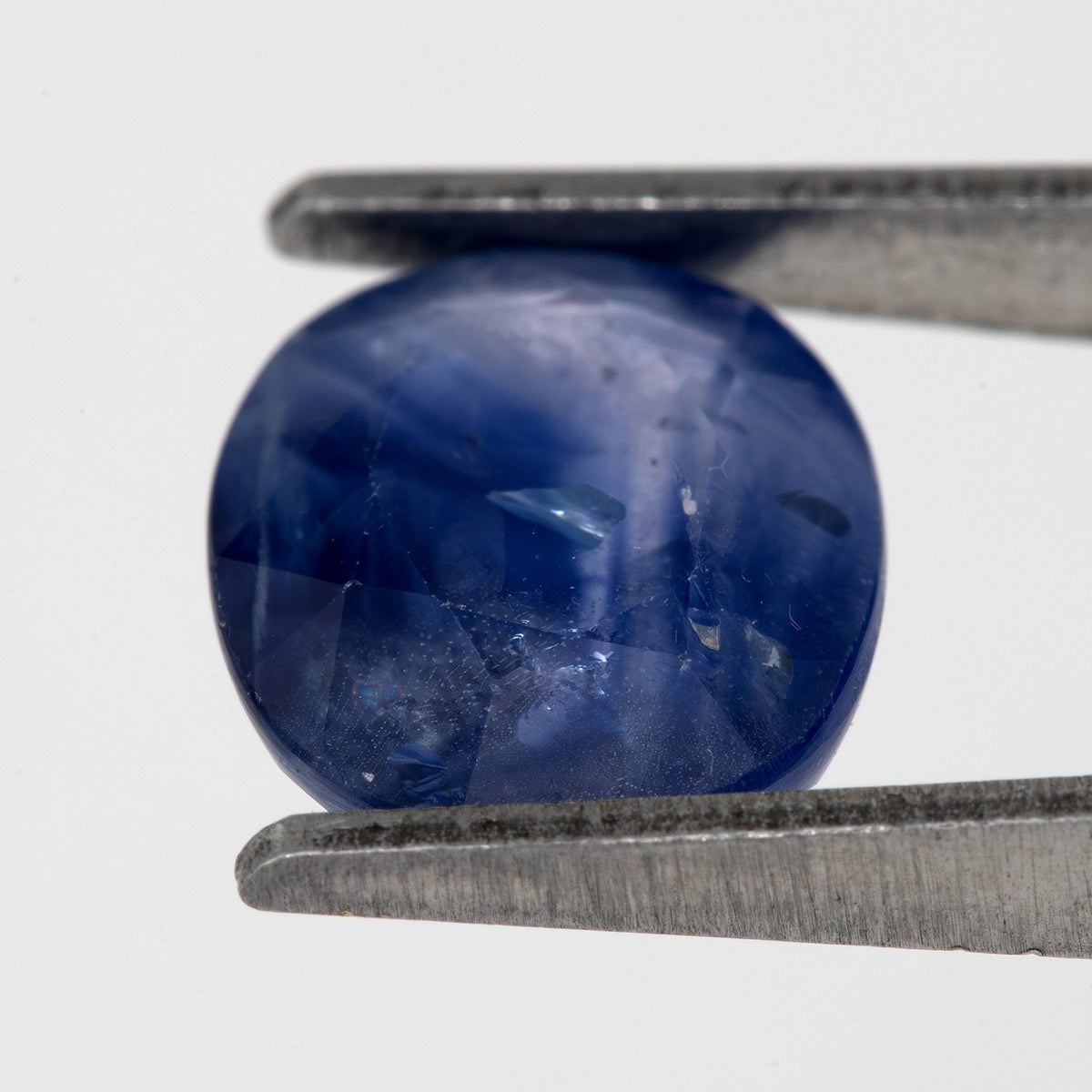 Tilted crown-side view of the oval blue sapphire in tweezers, showing a different pattern of facet reflections.