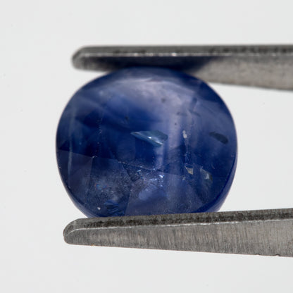 Tilted crown-side view of the oval blue sapphire in tweezers, showing a different pattern of facet reflections.